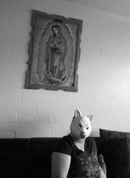 Black and White photo of woman with wolf mask sittin on couch with religious a painting hanging on wall