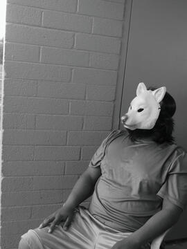 Black and White photo of a man with a wolf mask sitting outside in front of a brick wall